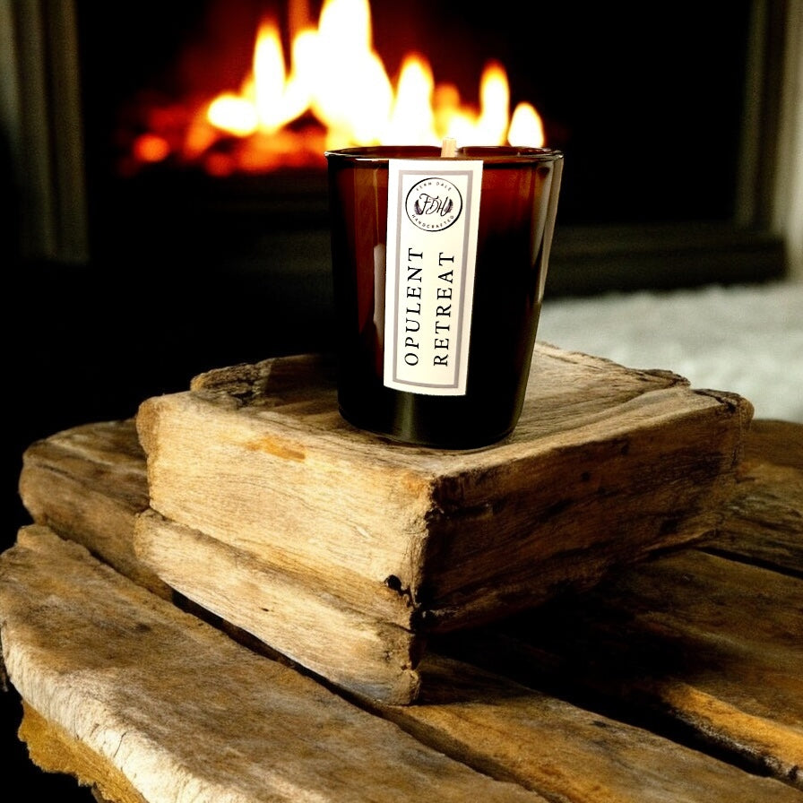 Opulent Retreat candle in a dark amber glass jar on stacked wooden blocks in front of a glowing fire.