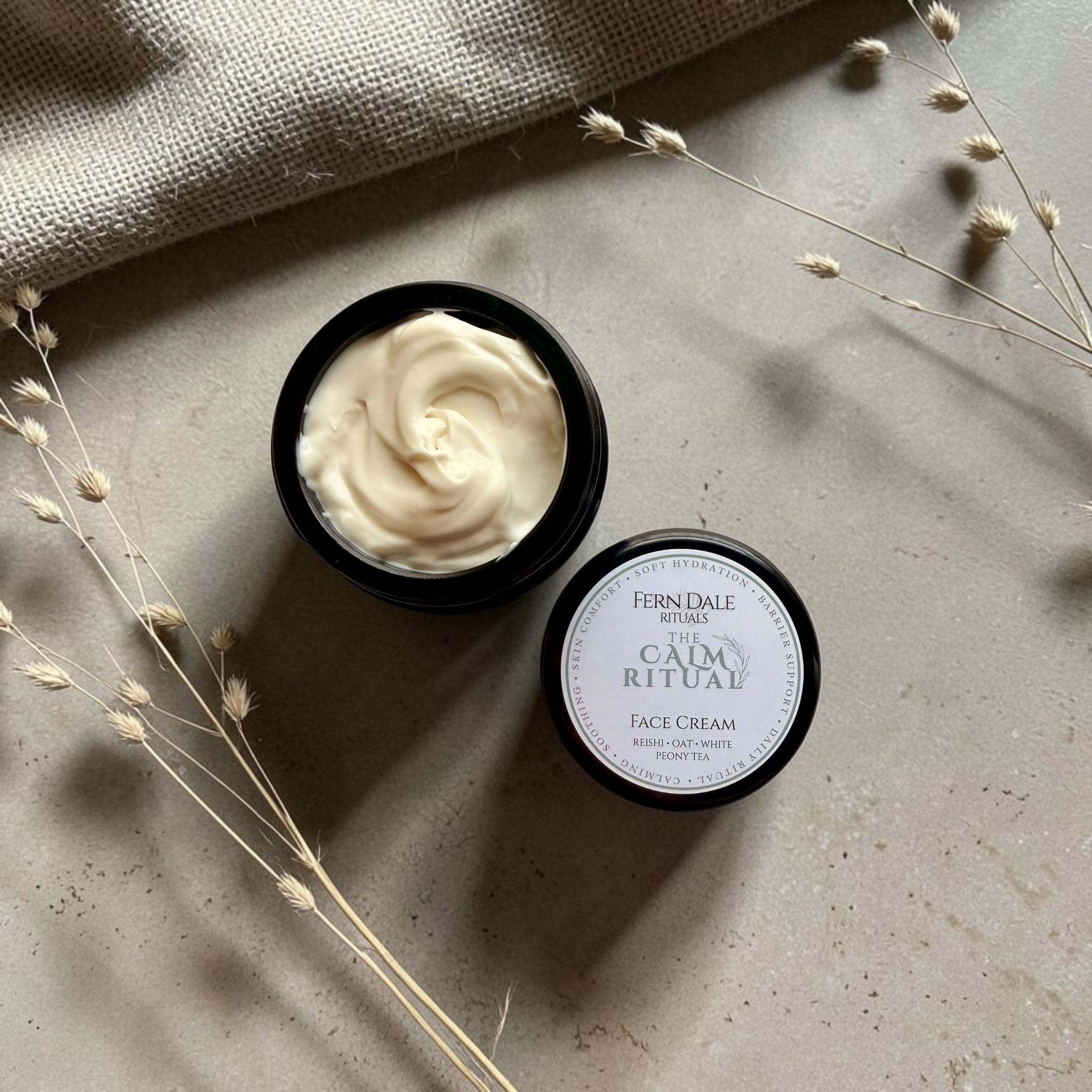The Calm Ritual - Face Cream in an open dark jar, with pale cream swirls, a labelled lid and dried grasses.